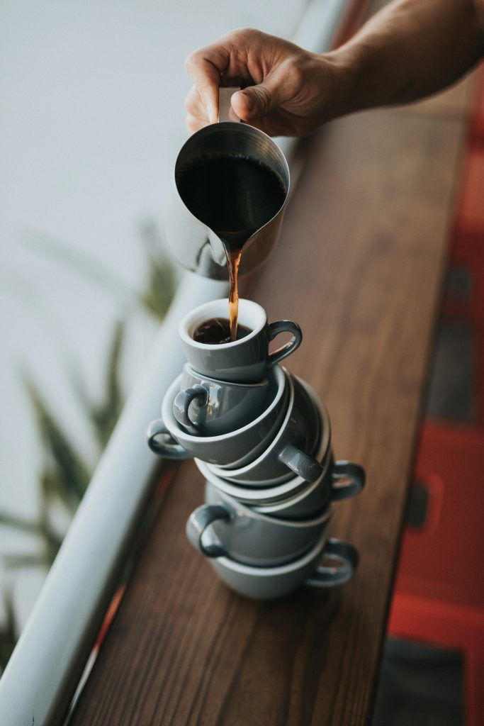 A precarious stack of coffee and espresso cup, with a brown hand holding a coffee pot, pouring into the top cup.