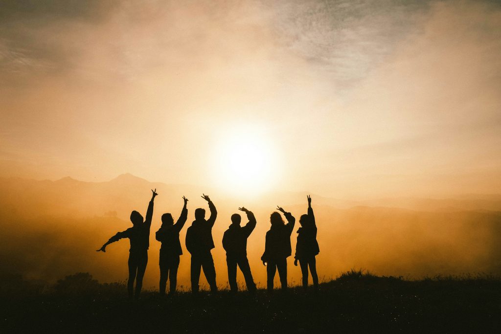 Silhouettes of a group of people standing on a hill at sunrise, raising their arms in celebration or greeting. The golden light and misty background create a hopeful, unified atmosphere.